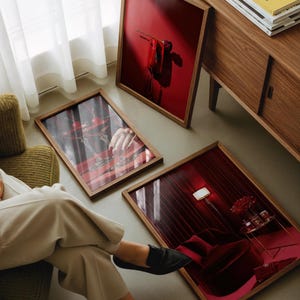 Peut inclure: Trois tirages d'art encadr&eacute;s avec des cadres en bois sont expos&eacute;s au sol. Les tirages pr&eacute;sentent des images rouges et roses, dont une nature morte avec des cerises, un rideau rouge et une main tenant un cocktail. Un meuble en bois et une chaise verte sont &eacute;galement visibles.