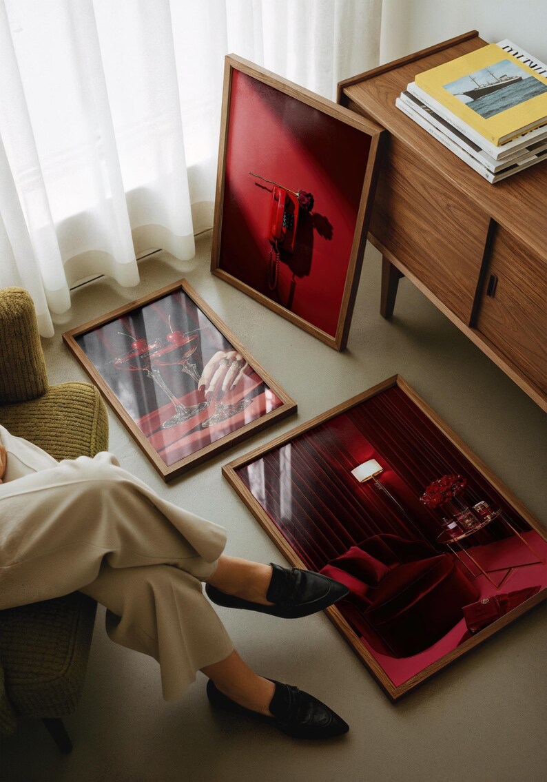 Peut inclure: Trois tirages d'art encadr&eacute;s avec des cadres en bois sont expos&eacute;s au sol. Les tirages pr&eacute;sentent des images rouges et roses, dont une nature morte avec des cerises, un rideau rouge et une main tenant un cocktail. Un meuble en bois et une chaise verte sont &eacute;galement visibles.