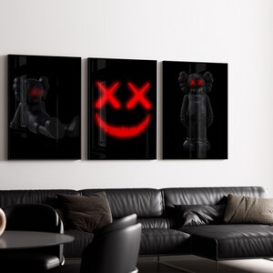 K&ouml;nnte beinhalten: Set aus drei schwarzen gerahmten Kunstdrucken mit stilisierten Figuren und einem roten Smiley-Gesicht. Die Drucke werden &uuml;ber einem schwarzen Ledersofa in einem modernen Wohnzimmer pr&auml;sentiert. Die Kunstwerke verleihen dem Raum eine moderne Note.