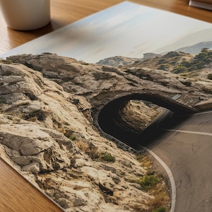 May include: A rectangular photographic print featuring a landscape scene of a paved road winding through a natural stone tunnel carved into a rugged, light brown rocky mountain side under a bright, clear sky with distant hills visible.