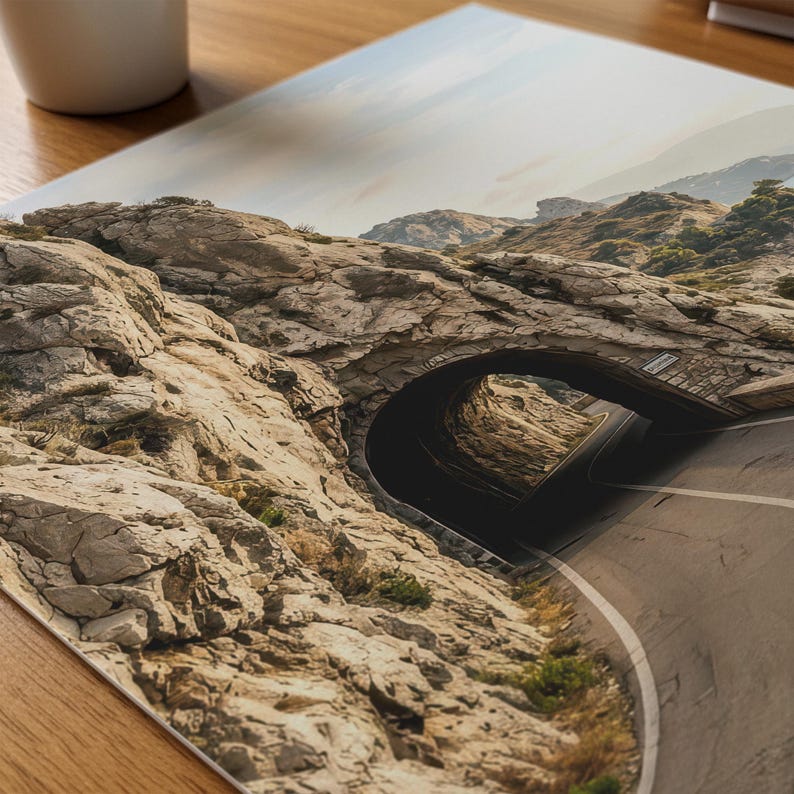 May include: A rectangular photographic print featuring a landscape scene of a paved road winding through a natural stone tunnel carved into a rugged, light brown rocky mountain side under a bright, clear sky with distant hills visible.
