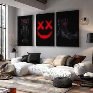 K&ouml;nnte beinhalten: Drei gerahmte schwarze Wandkunstwerke. Eines zeigt eine stehende schwarze Figur mit roten Augen, ein anderes ein rotes Neon-Smiley-Gesicht mit durchgestrichenen Augen und das dritte eine liegende schwarze Figur. Die Kunst h&auml;ngt &uuml;ber einem wei&szlig;en Ecksofa.
