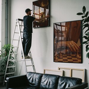 May include: A person on a ladder hangs framed art on a white wall. The art features a gold-toned trumpet and a hand playing chess. A black leather sofa and plants are in the foreground.