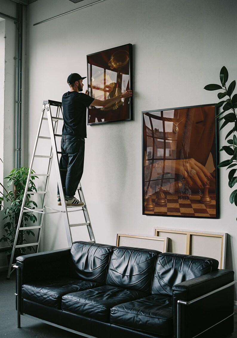 May include: A person on a ladder hangs framed art on a white wall. The art features a gold-toned trumpet and a hand playing chess. A black leather sofa and plants are in the foreground.