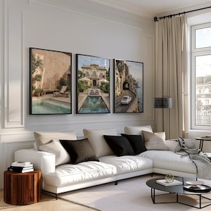 May include: Three framed wall art prints displayed above a white sectional sofa. The left print shows a pool and patio, the center print depicts a villa with a pool, and the right print features a white vintage car driving on a coastal road.
