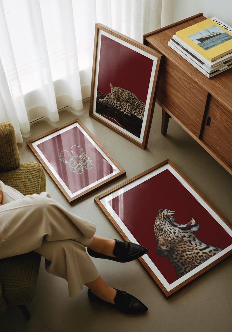 以下が含まれることがあります： 赤い背景と白い縁取りのある3つの額入りアートプリント。プリントにはヒョウが描かれており、1つは枝の上で寝ており、1つはあくびをしており、1つは靴のデザインです。フレームは茶色です。