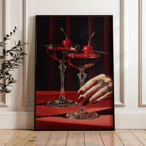 Peut inclure: Impression encadr&eacute;e de deux verres &agrave; pied remplis d'un liquide rouge, chacun garni d'une cerise. Une main aux ongles rouges et bagues touche un verre. Le fond est rouge fonc&eacute;.