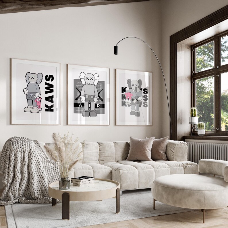 Puede incluir: Tres impresiones art&iacute;sticas enmarcadas con el personaje KAWS en varias poses. Las impresiones son en blanco, negro y gris, con la palabra "KAWS" en letras negras. La obra de arte se exhibe sobre un sof&aacute; beige.