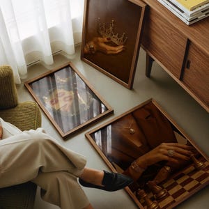 May include: Three framed artworks with wooden frames are displayed. One shows a hand holding a gold crown, another features red lips, and the third depicts a hand with jewellery and chess pieces. A wooden cabinet is in the background.