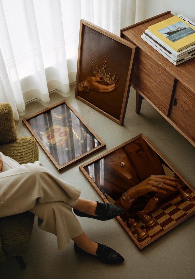 May include: Three framed artworks with wooden frames are displayed. One shows a hand holding a gold crown, another features red lips, and the third depicts a hand with jewellery and chess pieces. A wooden cabinet is in the background.