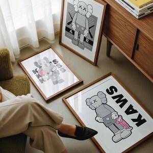 Puede incluir: Tres impresiones art&iacute;sticas enmarcadas con el personaje KAWS en varias poses y esquemas de color. Las impresiones est&aacute;n en marcos de madera y descansan en el suelo. Una impresi&oacute;n tiene la palabra "AIR" y otra la palabra "KAWS".