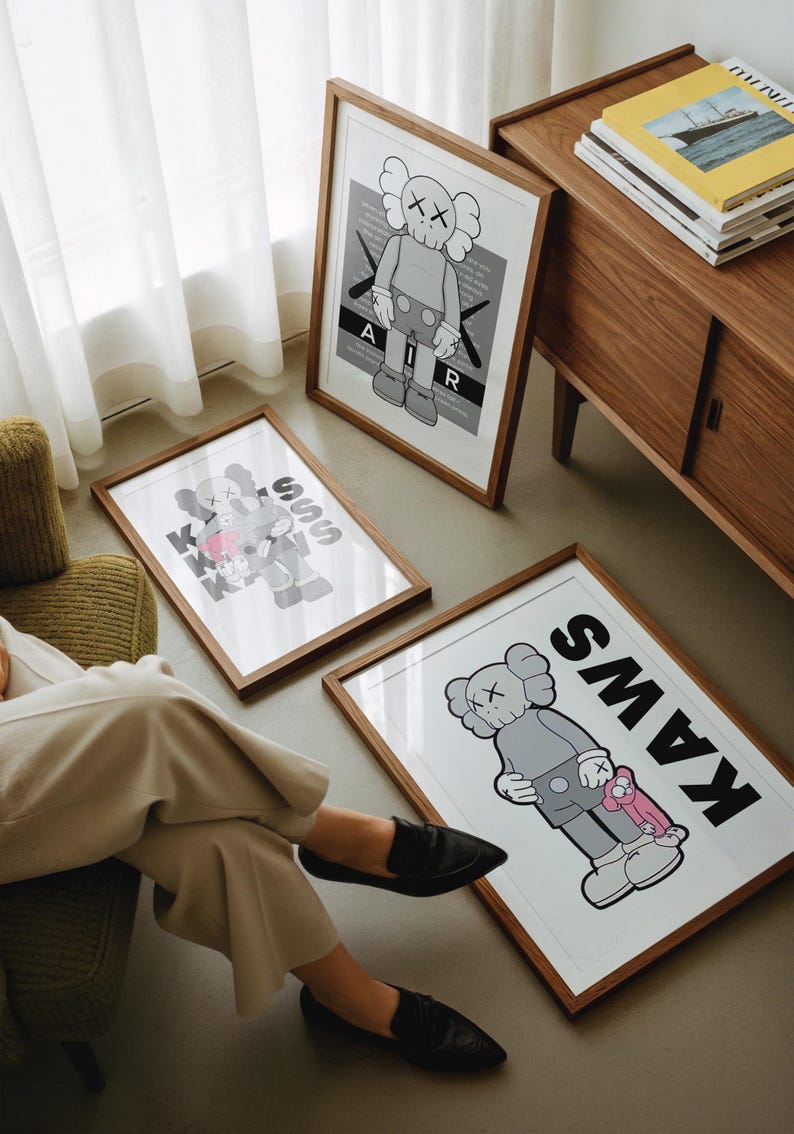 Puede incluir: Tres impresiones art&iacute;sticas enmarcadas con el personaje KAWS en varias poses y esquemas de color. Las impresiones est&aacute;n en marcos de madera y descansan en el suelo. Una impresi&oacute;n tiene la palabra "AIR" y otra la palabra "KAWS".