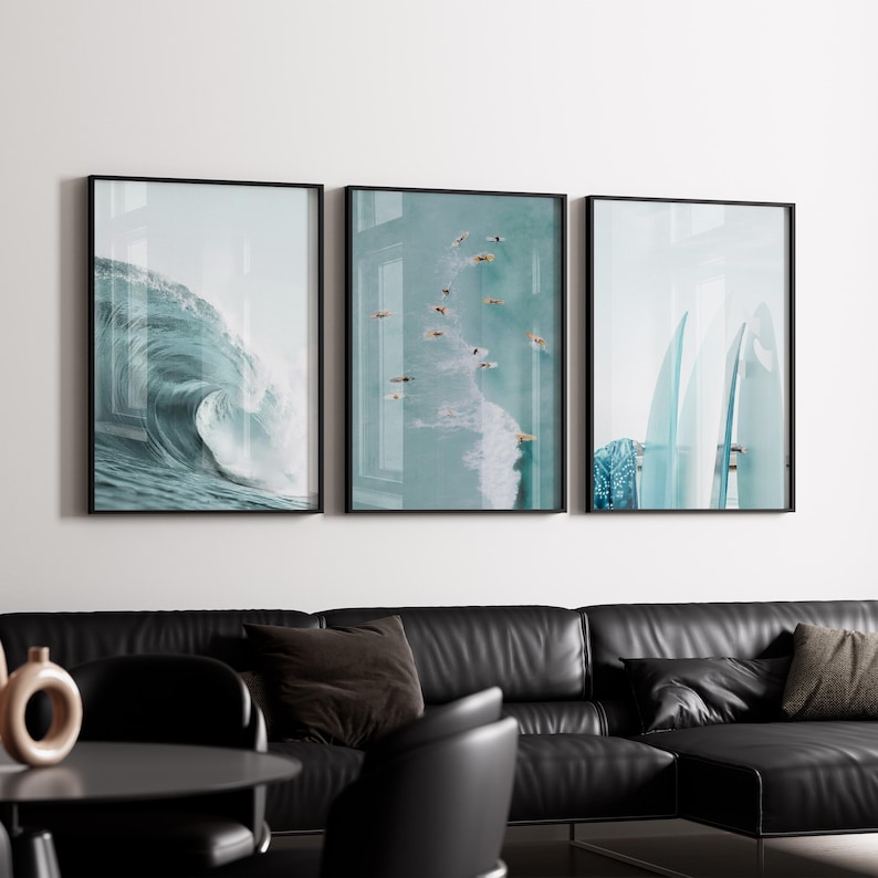 May include: Three framed art prints with black frames. The prints depict ocean scenes: a large wave, surfers in the water, and surfboards. The color palette is primarily shades of blue and white, creating a coastal theme.