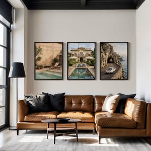 May include: A set of three framed wall art prints displayed above a brown leather sectional sofa. The prints feature a Mediterranean courtyard with a pool, a grand villa with a pool, and a white vintage car driving on a coastal road near a tunnel.
