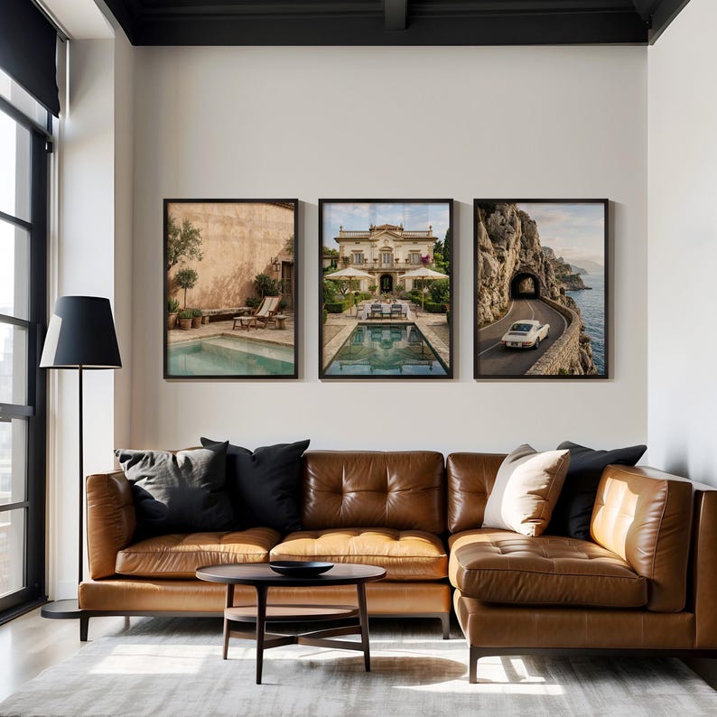 May include: A set of three framed wall art prints displayed above a brown leather sectional sofa. The prints feature a Mediterranean courtyard with a pool, a grand villa with a pool, and a white vintage car driving on a coastal road near a tunnel.