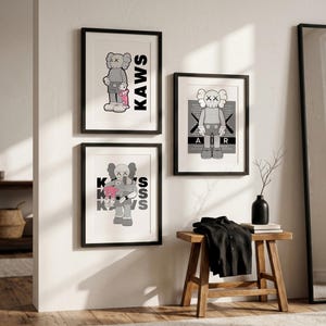 Puede incluir: Tres impresiones art&iacute;sticas enmarcadas con figuras de dibujos animados con ojos en forma de X. Las impresiones est&aacute;n en marcos negros y montadas en una pared blanca. Una impresi&oacute;n tiene la palabra "KAWS" en letras negras grandes. Las otras impresiones tienen las palabras "AIR" y "KAWS".