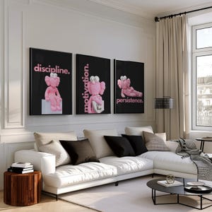 May include: Three framed art prints with black backgrounds and pink text. Each print features a pink cartoon figure and a motivational word: "discipline," "motivation," and "persistence." The prints are displayed above a white sectional sofa in a modern living room.