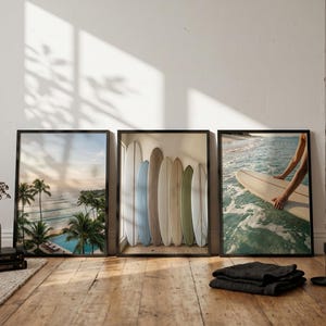 Op de afbeelding: Drie rechthoekige wandkunstprints in zwarte lijsten. De linker print toont een tropisch strand met palmbomen en een zwembad. De middelste print toont zes surfplanken tegen een muur. De rechter print toont een surfplank in de oceaan.