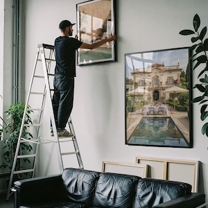 May include: A black leather sofa sits below two large framed wall art prints. One print depicts a grand villa with a swimming pool, while the other is being hung on the wall by a person standing on a tall silver metal step ladder.