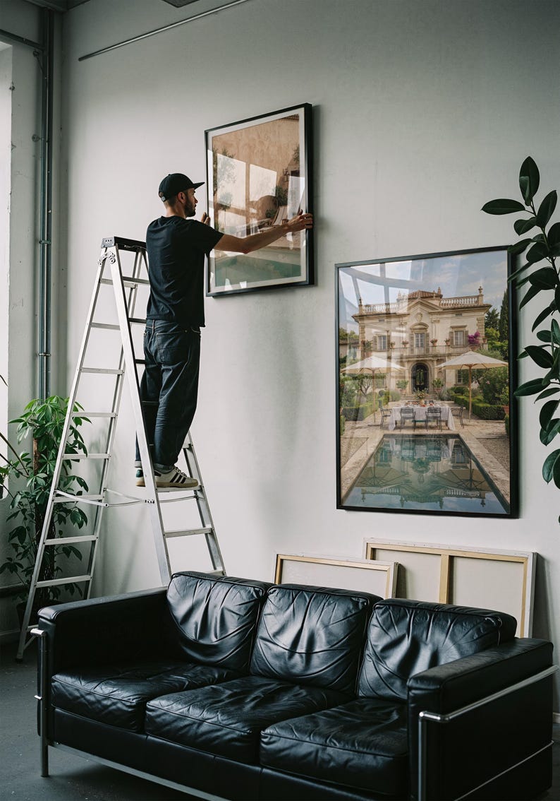 May include: A black leather sofa sits below two large framed wall art prints. One print depicts a grand villa with a swimming pool, while the other is being hung on the wall by a person standing on a tall silver metal step ladder.