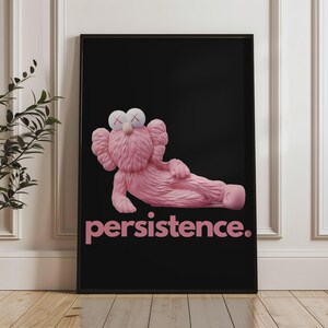May include: A framed art print featuring a pink cartoon figure with crossed-out eyes, reclining on a black background. The word "persistence" is printed in pink below the figure. The artwork is displayed against a white wall.