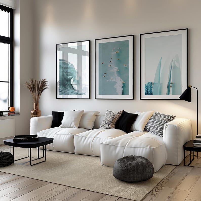 May include: A modern living room featuring a white sectional sofa with black and patterned throw pillows. Three framed ocean-themed art prints hang above the sofa. A black coffee table and a gray ottoman complete the decor.