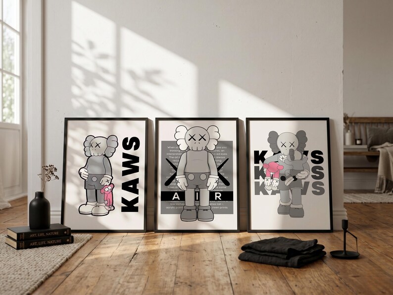 Puede incluir: Tres impresiones art&iacute;sticas enmarcadas con una figura de dibujos animados gris con ojos tachados. Las impresiones tienen marcos negros e incluyen las palabras "KAWS" y "AIR". Una impresi&oacute;n muestra la figura sosteniendo un personaje rosa. Las impresiones se exhiben sobre un suelo de madera.