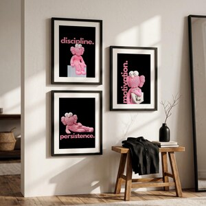 May include: Three framed art prints with black frames hang on a white wall. Each print features a pink cartoon character on a black background with a motivational word in pink: "discipline," "motivation," and "persistence."