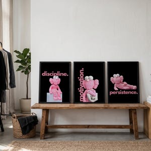May include: Three framed art prints featuring pink cartoon figures on a black background. Each print has a different word in pink text: "discipline.", "motivation.", and "persistence.". The prints are displayed on a wooden bench.