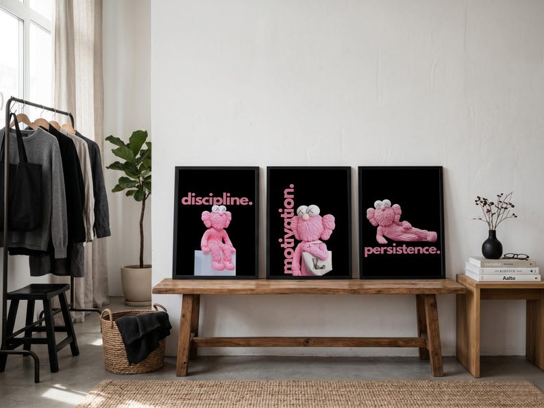 May include: Three framed art prints featuring pink cartoon figures on a black background. Each print has a different word in pink text: "discipline.", "motivation.", and "persistence.". The prints are displayed on a wooden bench.