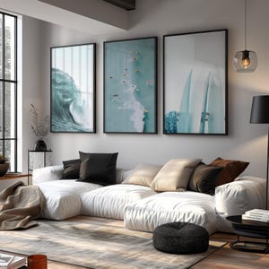 May include: Three framed wall art pieces with black frames. The art features ocean themes: a crashing wave, surfers in the water, and surfboards. The art hangs above a white sectional sofa with black and tan pillows.