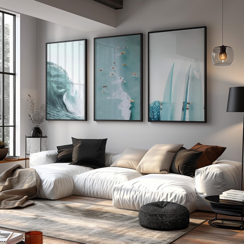 May include: Three framed wall art pieces with black frames. The art features ocean themes: a crashing wave, surfers in the water, and surfboards. The art hangs above a white sectional sofa with black and tan pillows.