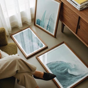 May include: Three framed art prints with wooden frames are displayed on the floor and leaning against a wall. The prints depict ocean waves and surfboards. A wooden cabinet and a person wearing black shoes are also visible.