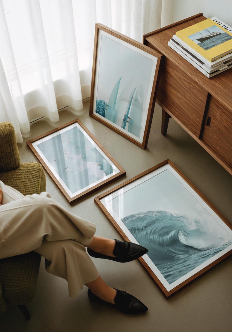 May include: Three framed art prints with wooden frames are displayed on the floor and leaning against a wall. The prints depict ocean waves and surfboards. A wooden cabinet and a person wearing black shoes are also visible.