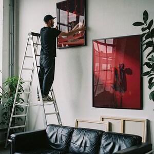 Peut inclure: Une personne sur une &eacute;chelle accroche une &oelig;uvre d'art encadr&eacute;e sur un mur blanc. L'&oelig;uvre pr&eacute;sente un design abstrait rouge et noir. Un canap&eacute; en cuir noir est en dessous. D'autres &oelig;uvres encadr&eacute;es sont visibles.