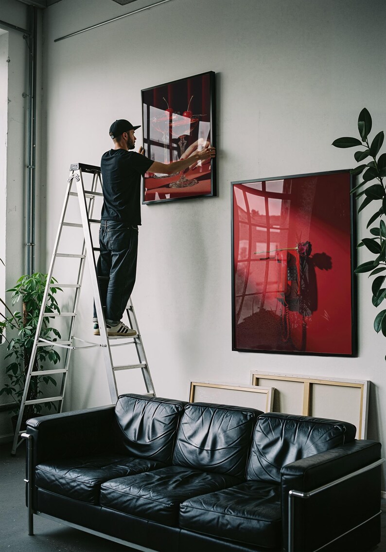 Peut inclure: Une personne sur une &eacute;chelle accroche une &oelig;uvre d'art encadr&eacute;e sur un mur blanc. L'&oelig;uvre pr&eacute;sente un design abstrait rouge et noir. Un canap&eacute; en cuir noir est en dessous. D'autres &oelig;uvres encadr&eacute;es sont visibles.