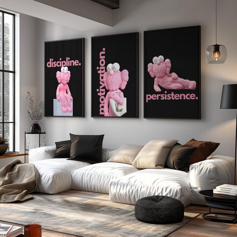 May include: Three framed art prints with black backgrounds and pink text. Each features a pink cartoon figure in different poses. The text reads "discipline.", "motivation.", and "persistence." The art hangs above a white sectional sofa.