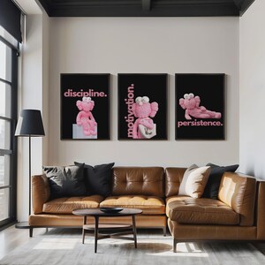 May include: Three framed art prints with black backgrounds and pink text. Each print features a pink cartoon character in different poses, with the words "discipline," "motivation," and "persistence." The prints hang above a brown leather sectional sofa.