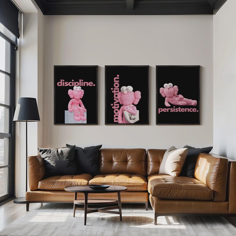 May include: Three framed art prints with black backgrounds and pink text. Each print features a pink cartoon character in different poses, with the words "discipline," "motivation," and "persistence." The prints hang above a brown leather sectional sofa.
