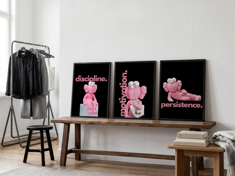 May include: Three framed art prints with black backgrounds and pink text. The prints feature a pink cartoon character in different poses, with the words "discipline," "motivation," and "persistence." The prints are displayed on a wooden bench.
