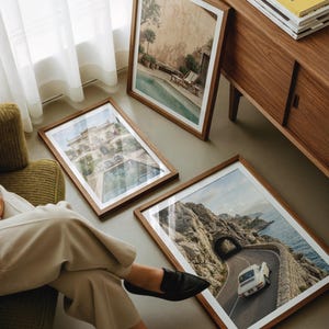 May include: Three rectangular wooden frames containing photographic prints. One print shows a swimming pool, another depicts a large villa with a pool, and the third features a white car driving on a coastal road through a tunnel.
