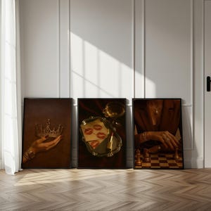 May include: Three framed art prints leaning against a white wall. The prints feature a hand holding a crown, a tray with lipstick and kisses, and a person playing chess. The prints have a brown and gold colour scheme.