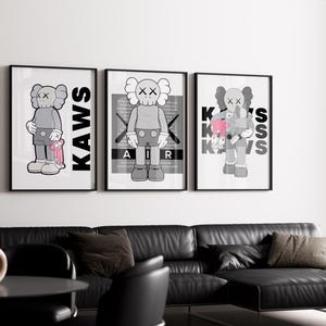 Puede incluir: Tres impresiones art&iacute;sticas enmarcadas con una figura de dibujos animados gris con ojos en forma de X. Las impresiones tienen marcos negros y las palabras "KAWS" o "AIR" en negrita. La obra de arte se exhibe sobre un sof&aacute; de cuero negro.