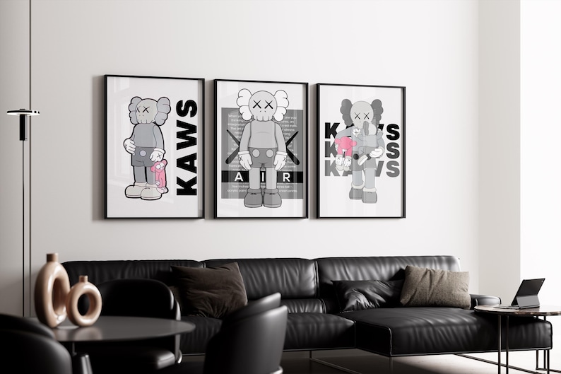 Puede incluir: Tres impresiones art&iacute;sticas enmarcadas con una figura de dibujos animados gris con ojos en forma de X. Las impresiones tienen marcos negros y las palabras "KAWS" o "AIR" en negrita. La obra de arte se exhibe sobre un sof&aacute; de cuero negro.