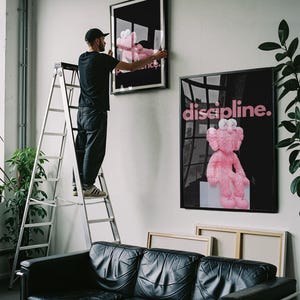 May include: A person hangs framed art on a white wall. The art features a pink plush toy on a black background. The word "discipline" is written in pink on one piece. A black leather sofa sits below the art.