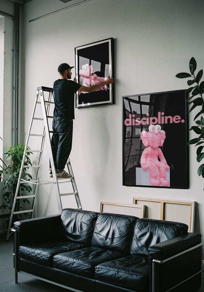 May include: A person hangs framed art on a white wall. The art features a pink plush toy on a black background. The word "discipline" is written in pink on one piece. A black leather sofa sits below the art.