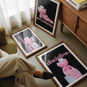 May include: Three framed art prints with a black background and pink text. Each print features a pink cartoon figure in a different pose. The text on the prints reads "persistence", "motivation", and "discipline". The frames are brown.