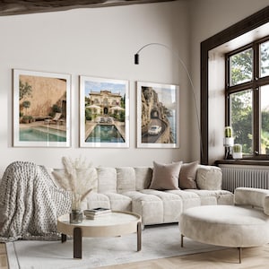May include: Three framed wall art prints displayed above a beige modular sofa. The left print shows a pool with a lounge chair, the center print depicts a villa with a pool, and the right print features a white car driving on a coastal road.