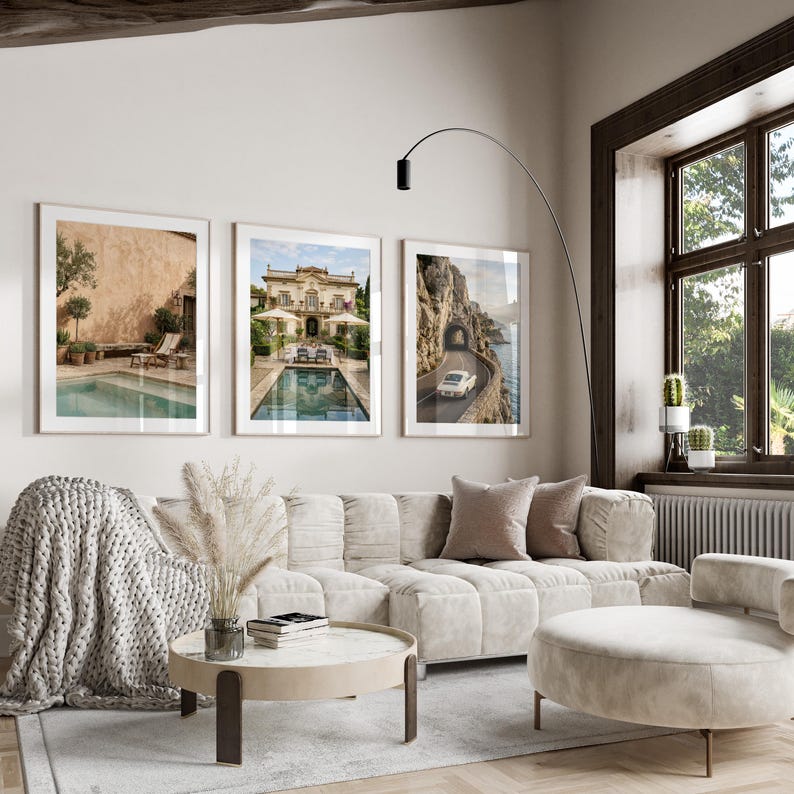 May include: Three framed wall art prints displayed above a beige modular sofa. The left print shows a pool with a lounge chair, the center print depicts a villa with a pool, and the right print features a white car driving on a coastal road.