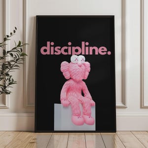 May include: Framed art print featuring a pink, furry character with crossed-out eyes, seated on a gray block against a black background. The word "discipline" is displayed in pink above the character.